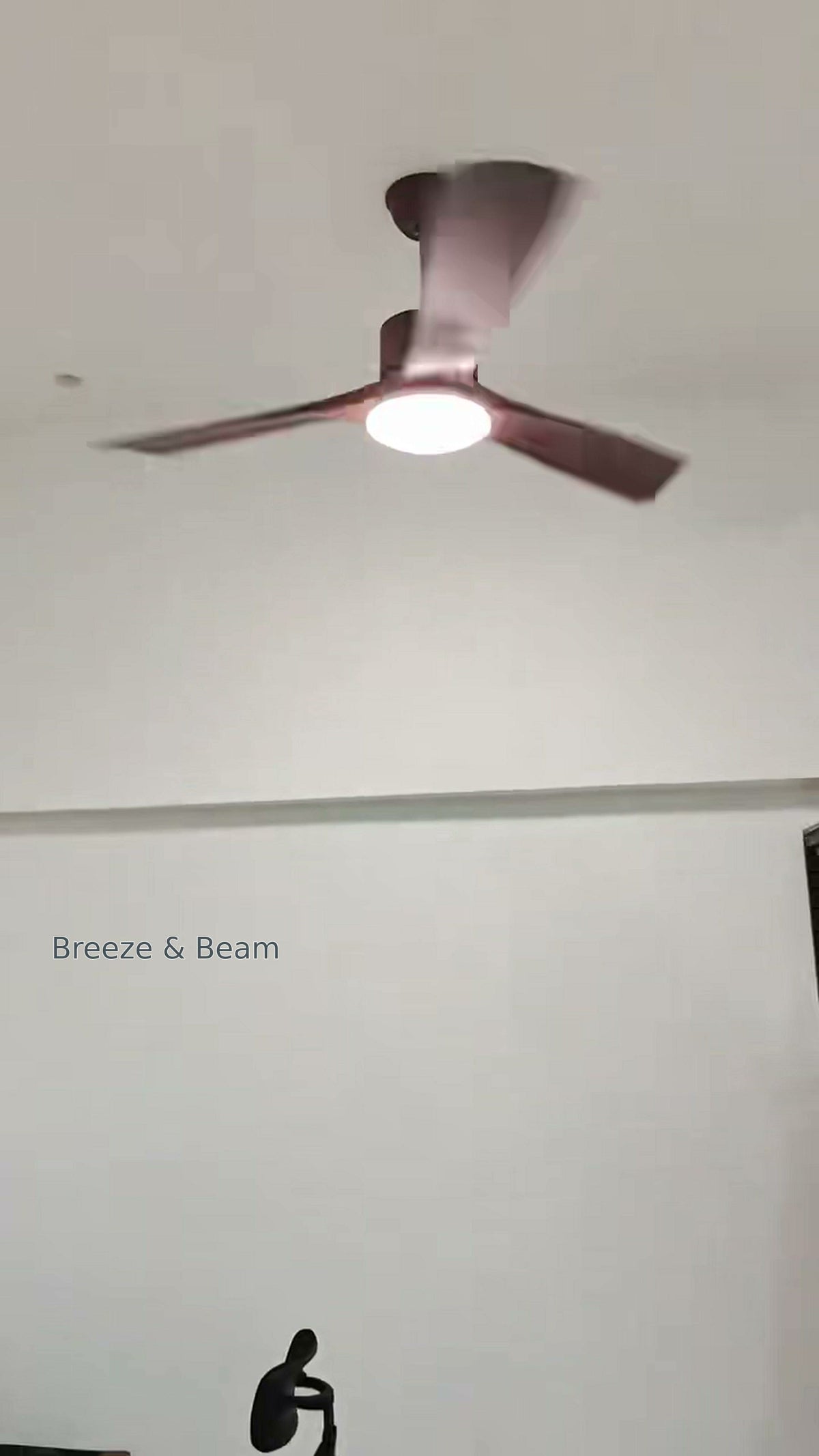 Three warm brown ceiling fan blades around an illuminated round white light against a white ceiling, with a dark office chair below.